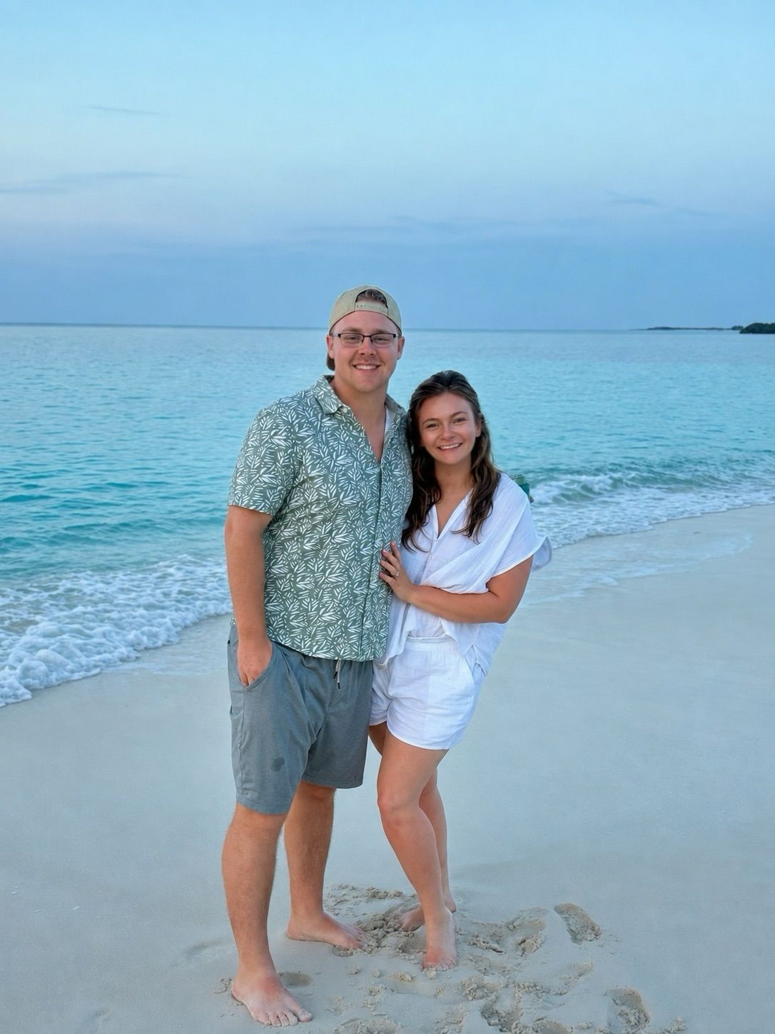 Two people smiling and standing barefoot on a sandy beach at sunset by calm turquoise water
