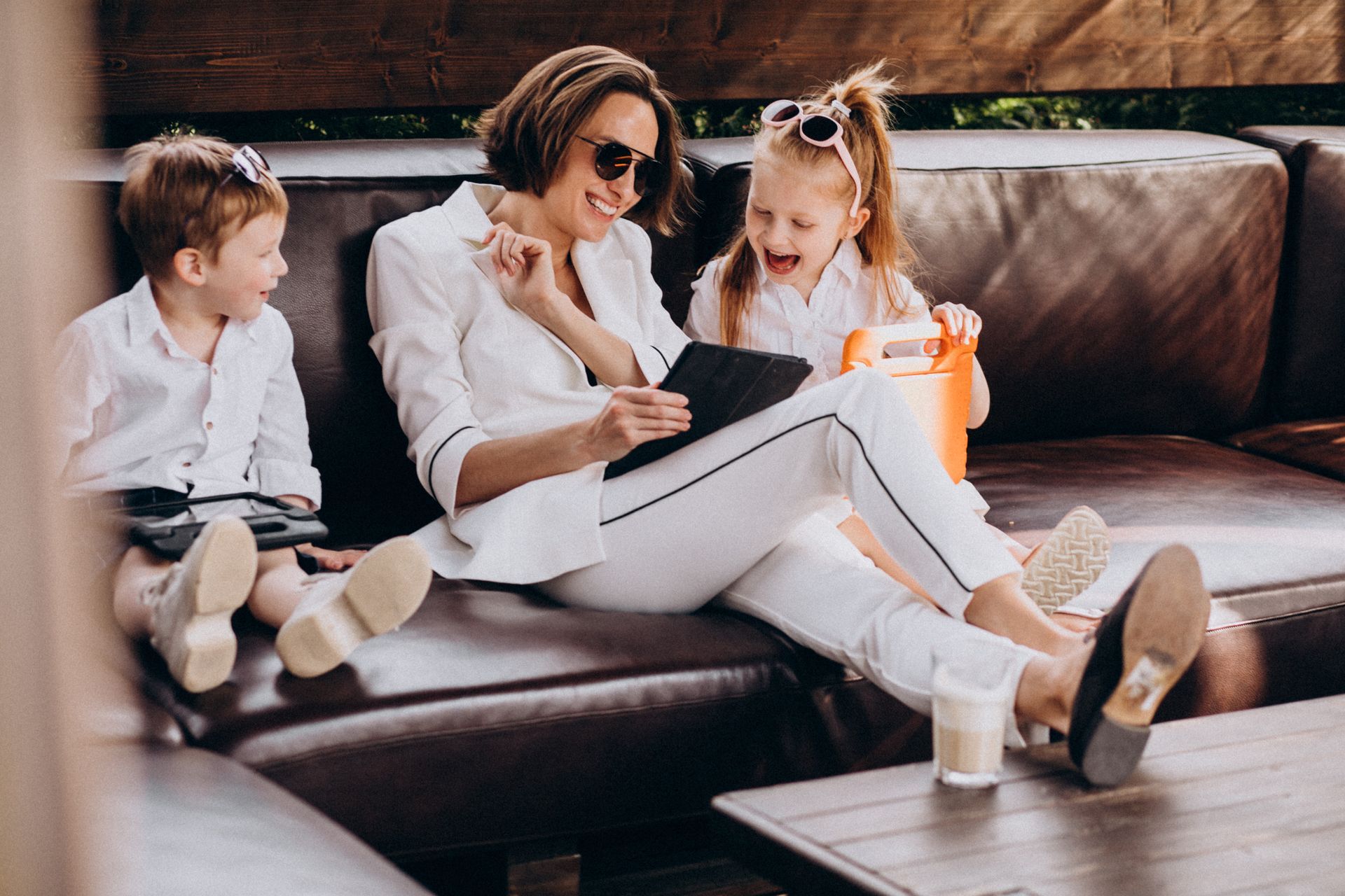 Three people sitting on a brown leather couch, sharing a tablet in a cozy living room.