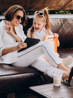 Two women in white outfits smiling on a couch, one holding a tablet, with a small dog nearby.