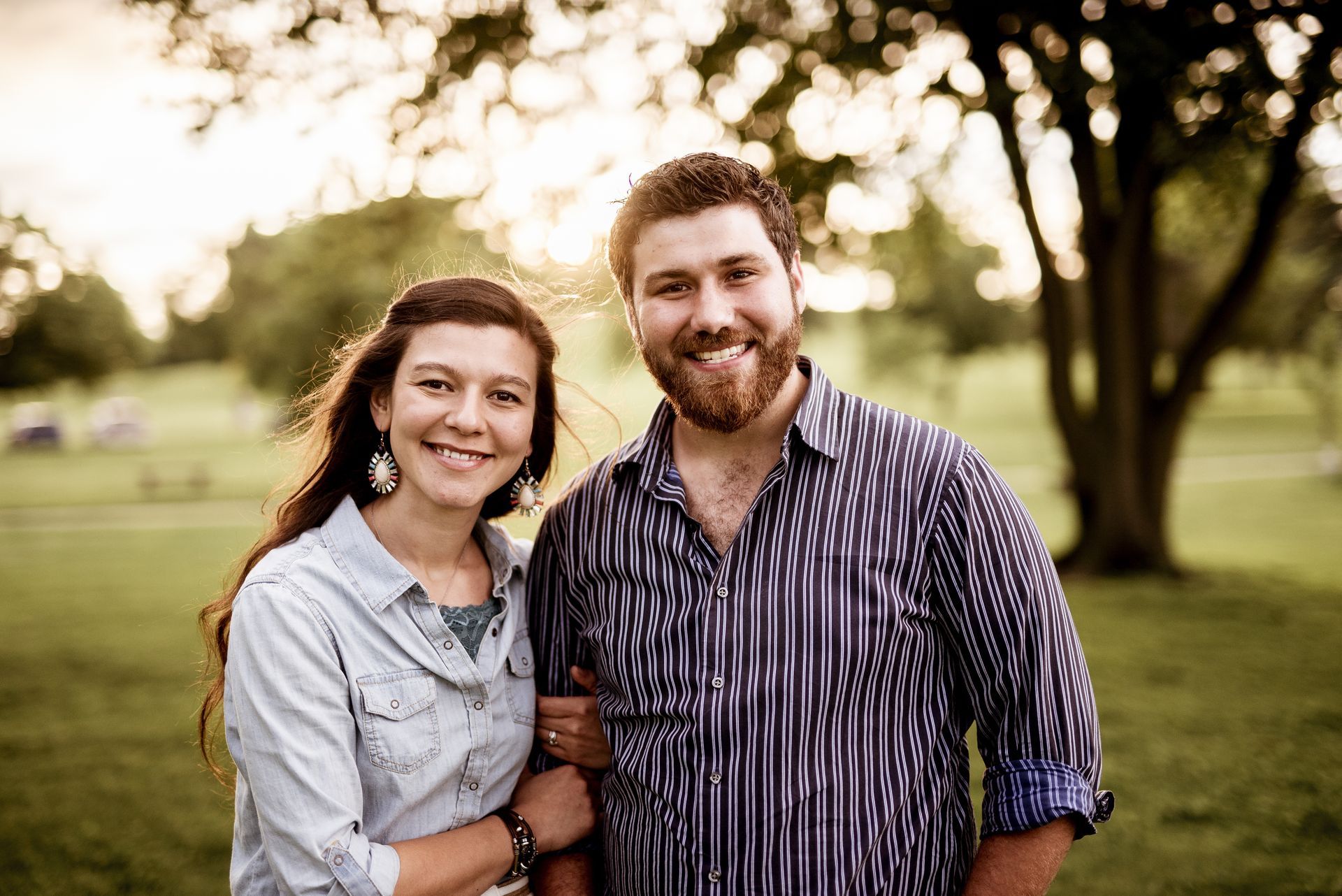 Two smiling people standing arm in arm in a sunlit park with trees in the background