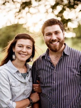 Smiling couple standing outdoors, arm in arm, with soft trees in the background