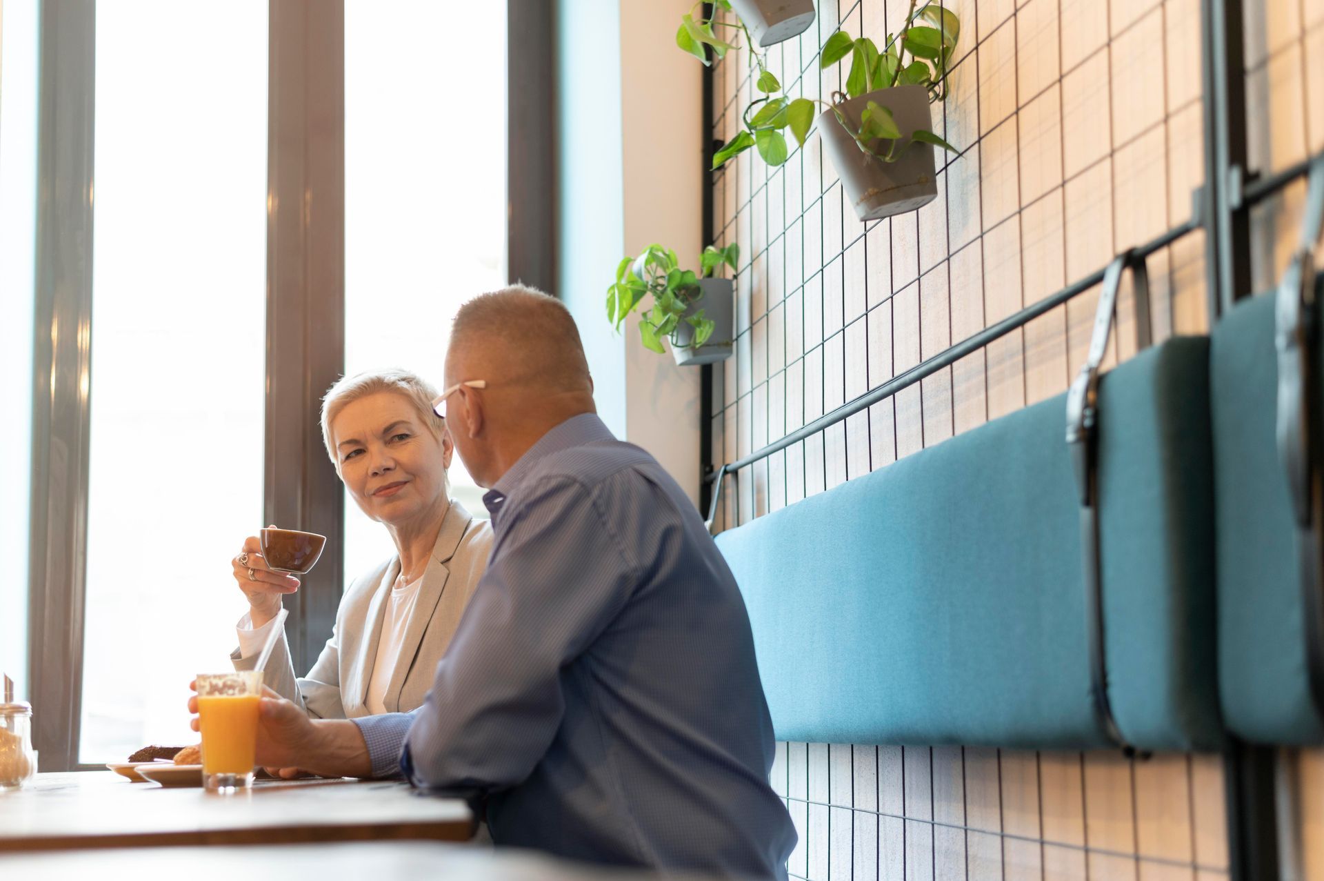Two people chatting at a cafe table beside blue seating and a wall grid with hanging plants.