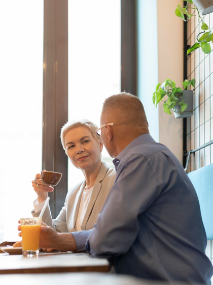 Two people chatting at a café table, raising drinks by a bright window