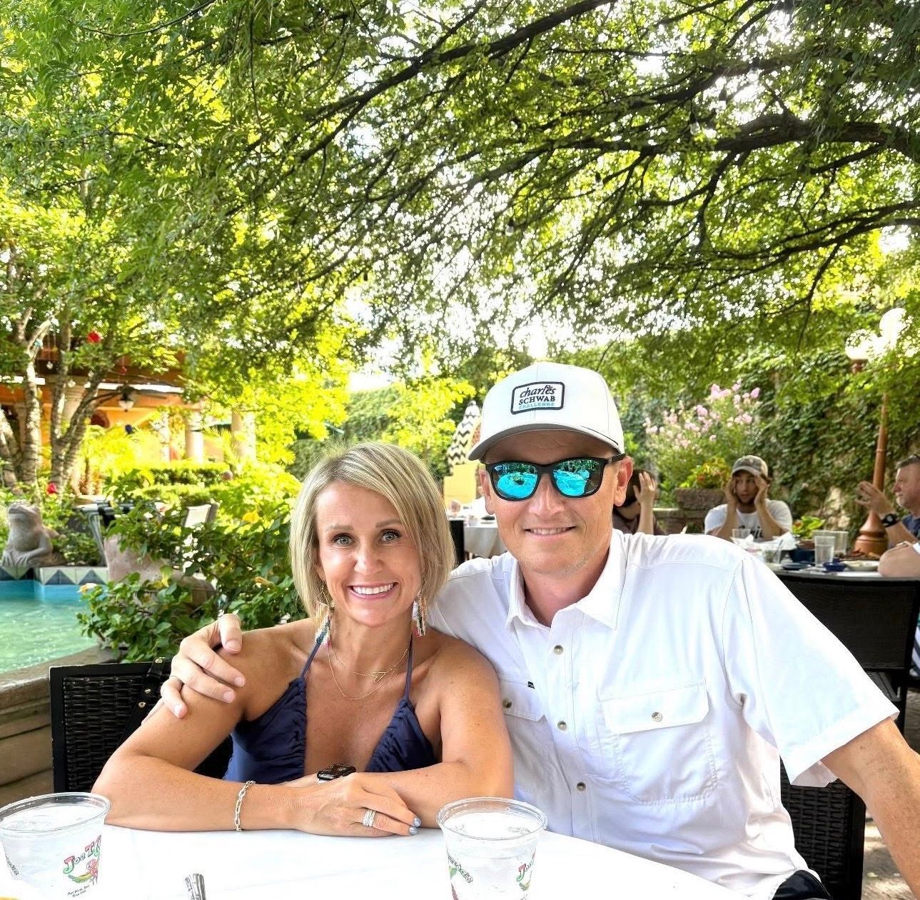 Couple smiling at an outdoor café under leafy trees, with drinks on the table and a pool in the background
