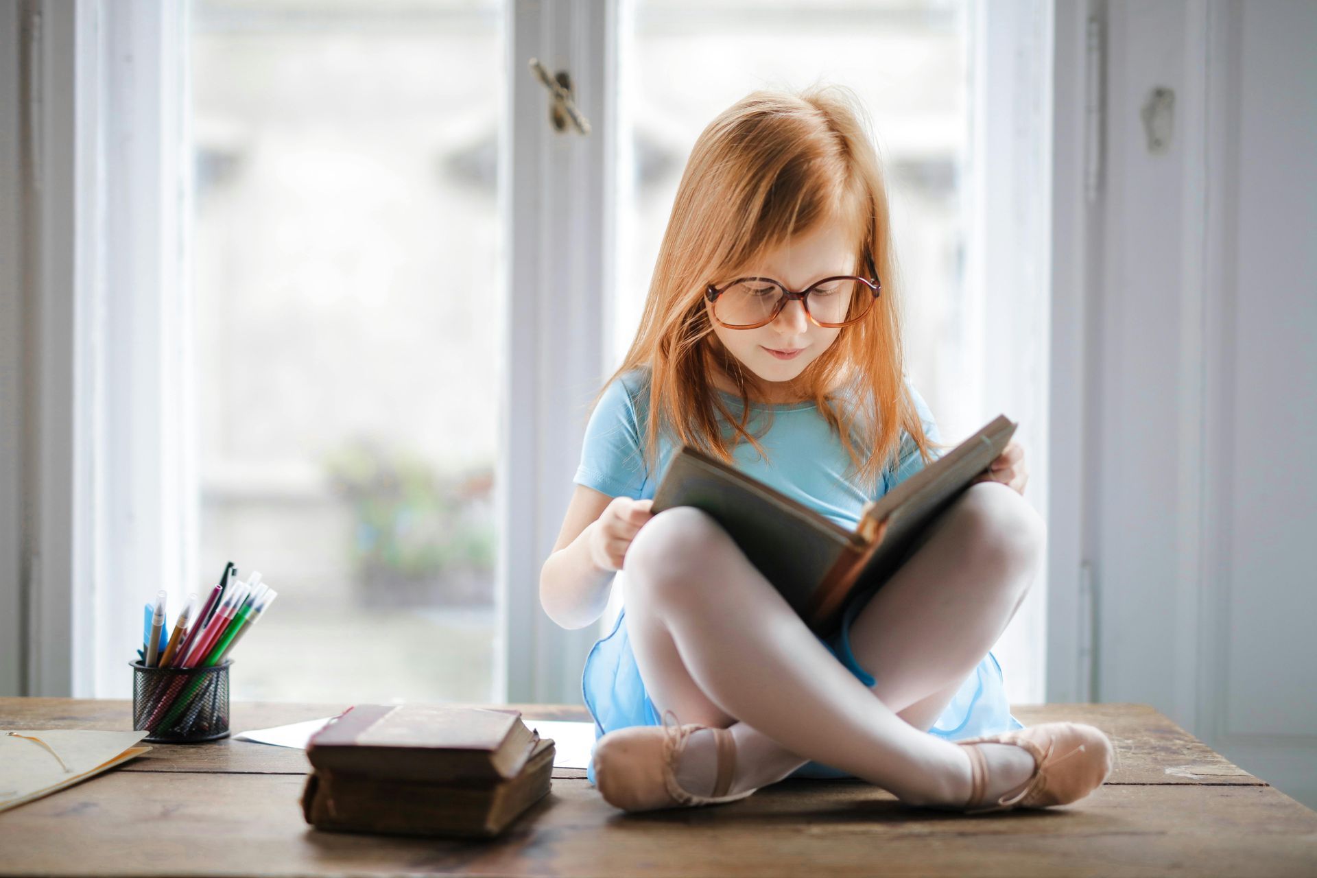 Jong kind met bril leest een boek bij een licht raam, zittend aan een houten tafel met boeken en potloden in de buurt.