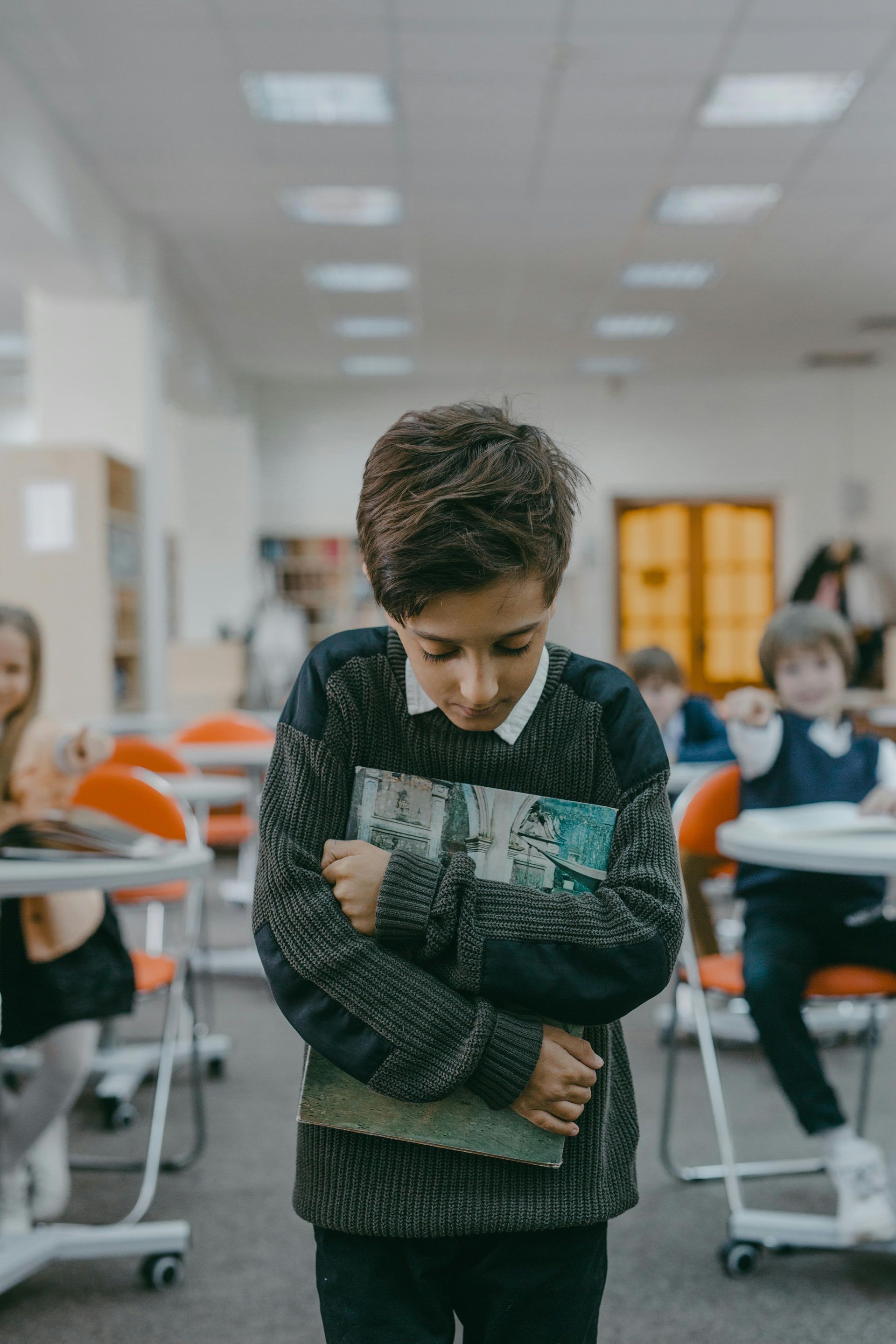 Een leerling staat met de armen over elkaar in een klaslokaal en kijkt naar beneden, met bureaus en zittende klasgenoten op de achtergrond.