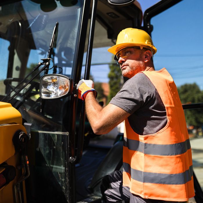 A construction worker in a yellow hard hat and safety vest gets ready to operate a yellow excavator