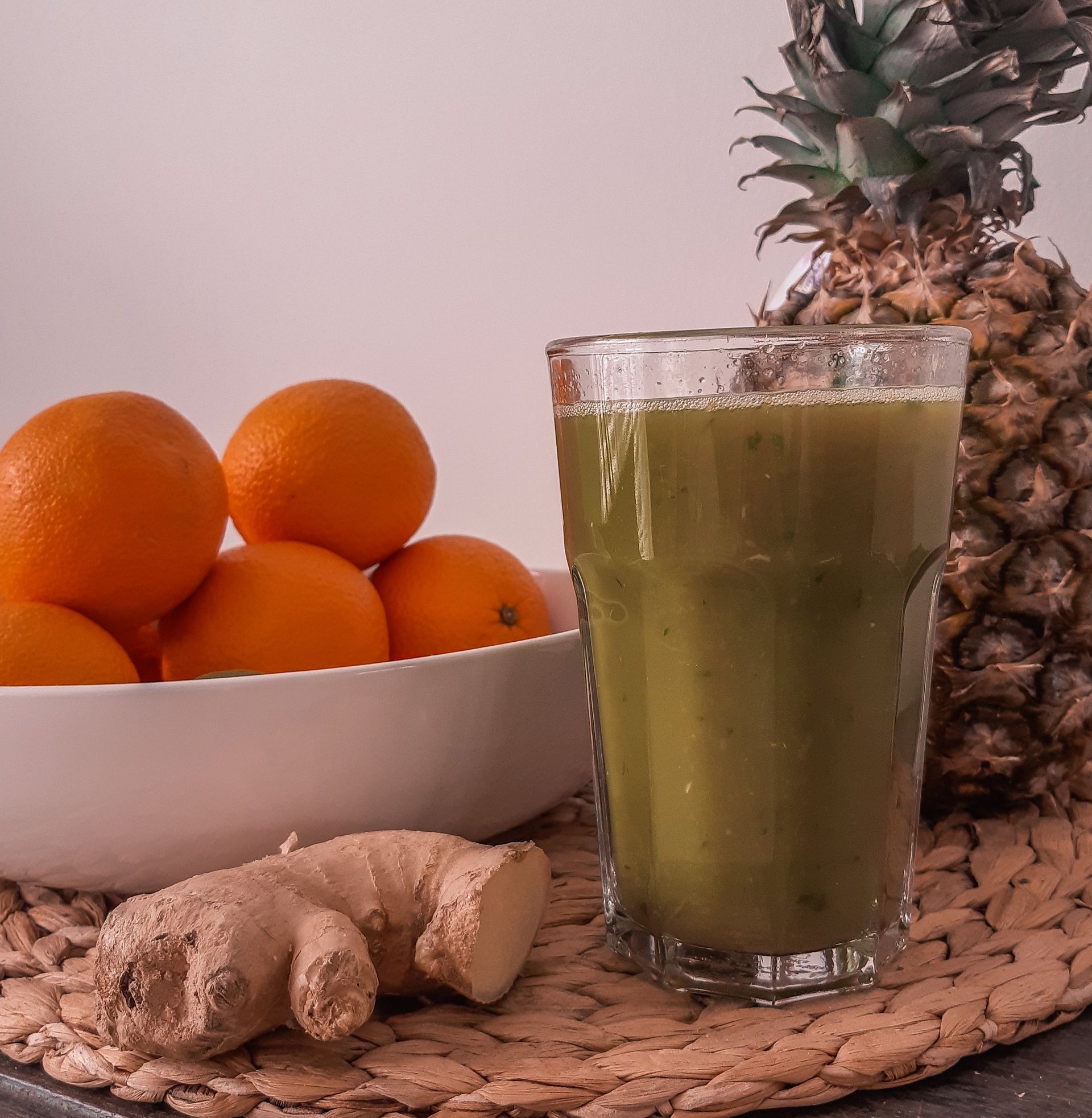 Een groene smoothie in een glas, naast sinaasappels, gemberwortel en een ananas op een geweven mat.