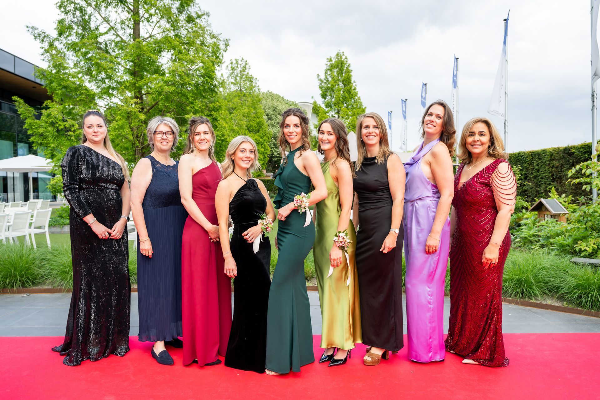 Een groep vrouwen in kleurrijke avondjurken poseert op een rode loper in de buitenlucht.