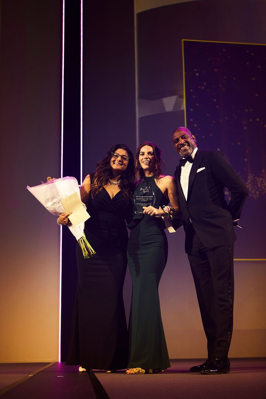 Drie personen in galakleding poseren op het podium met een boeket en een trofee onder paarse verlichting.