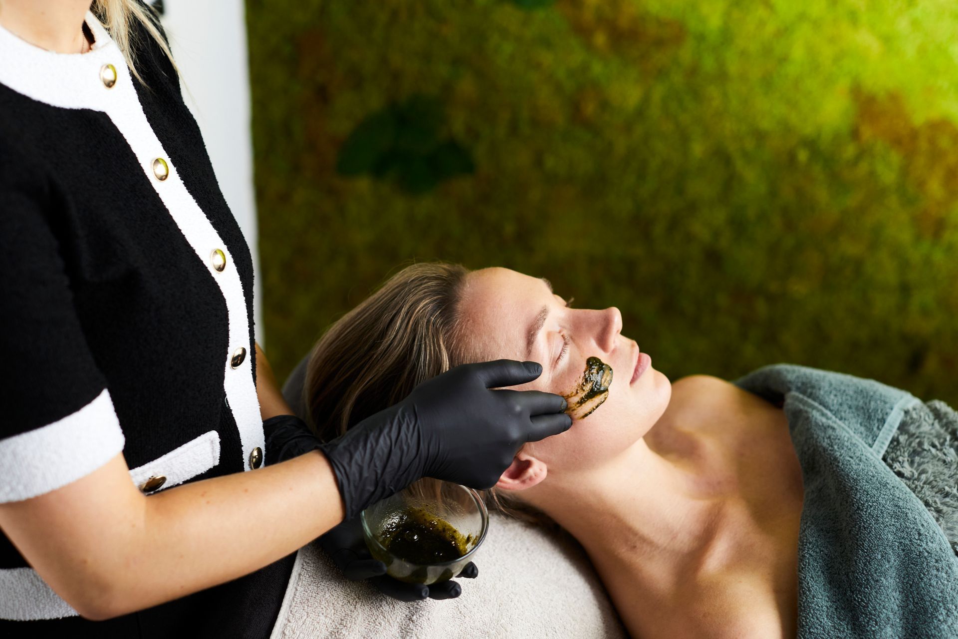 Persoon met een groen gezichtsmasker en een grijze hoofddoek, liggend in een spa-achtige omgeving.