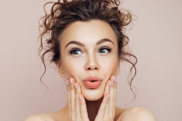 Vrouw met krullend haar raakt haar wangen aan en kijkt verbaasd tegen een zachtroze achtergrond.