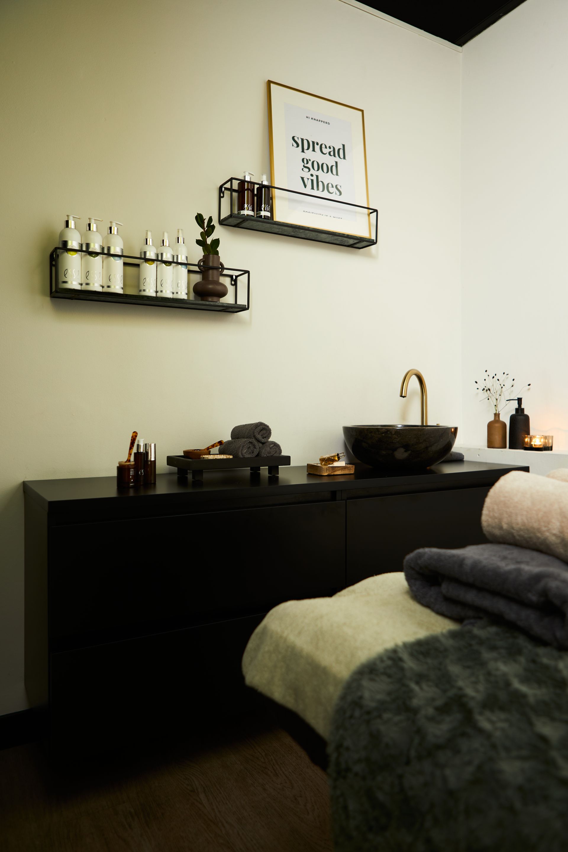 Minimalistische spa-behandelkamer met zwart aanrechtblad, gouden kraan en massagetafel met opgevouwen handdoeken.