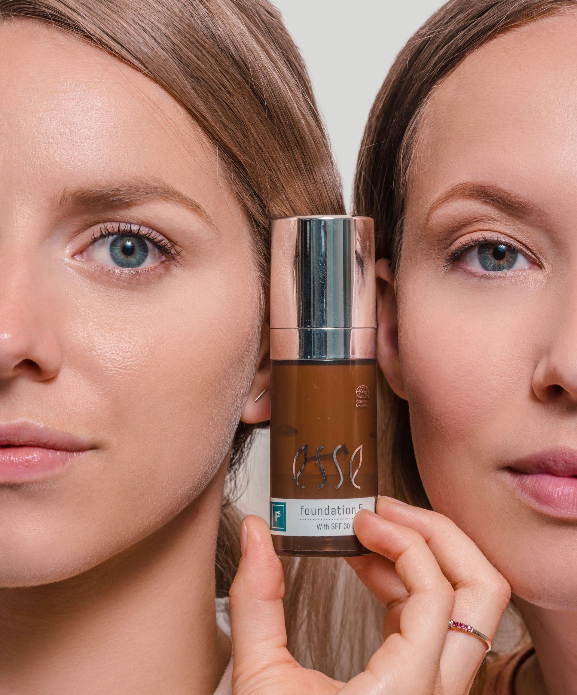 Twee vrouwen houden een bruine fles huidverzorgingsproducten met zilveren dop vast, tegen een roze achtergrond.