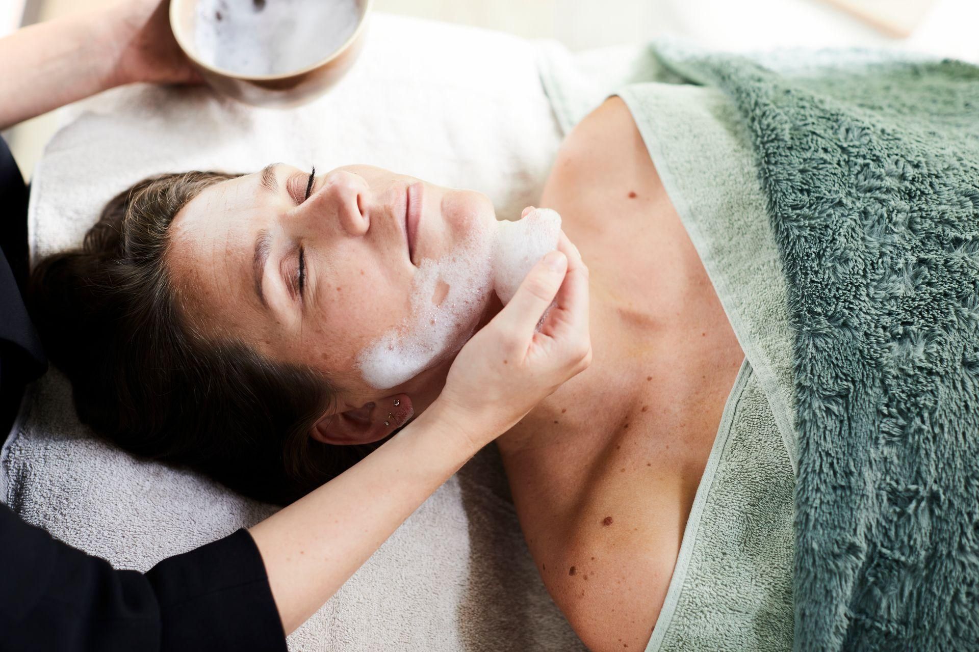 Persoon ondergaat een gezichtsbehandeling met witte crème terwijl ze op een massagetafel ligt.