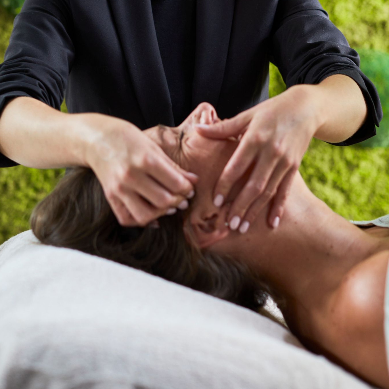 Een massagetherapeut geeft een hoofdmassage aan een cliënt die buiten op een witte handdoek ligt.