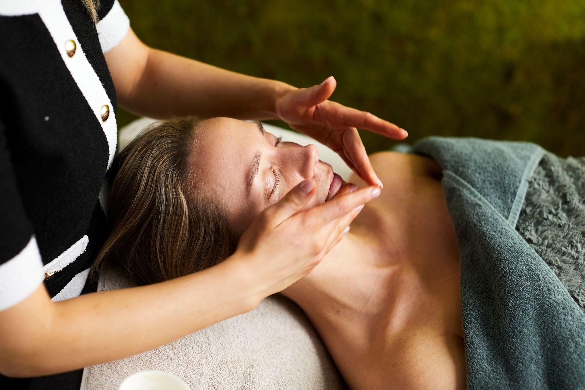 Een spa-therapeut geeft een gezichtsmassage aan een cliënt die onder een grijze handdoek ligt.