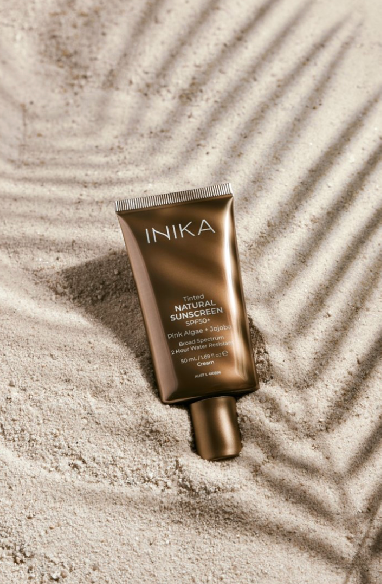 Bronzen INIKA-buislamp op beige zand met zachte schaduwen van palmbladeren.
