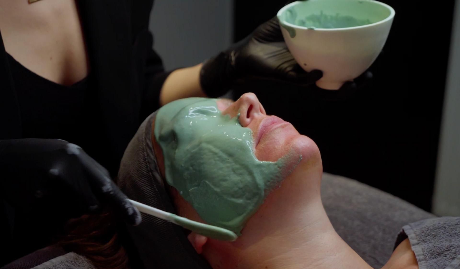 Een persoon krijgt een groen gezichtsmasker in een spa, aangebracht met een kom en een kwastje op het gezicht.