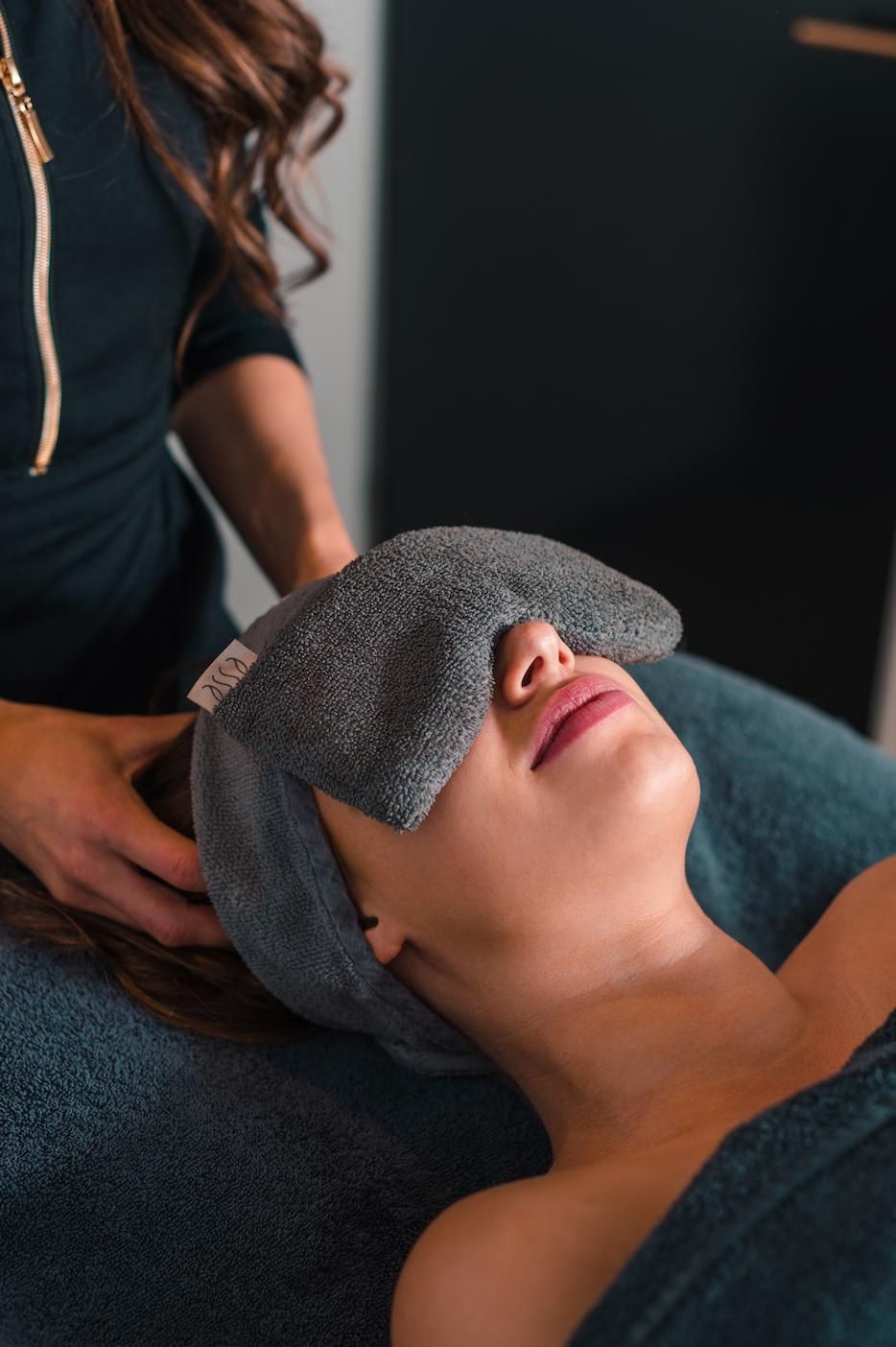 Een spa-therapeut legt een warme handdoek over de ogen van een liggende cliënt tijdens een gezichtsbehandeling.
