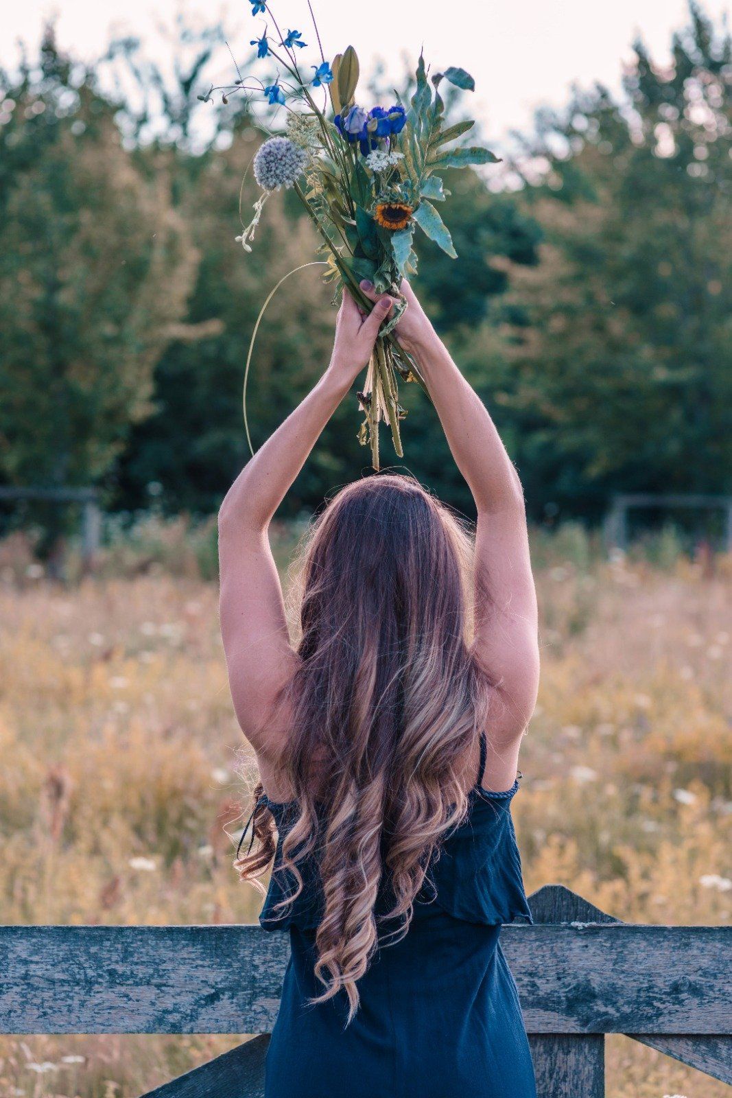 Vrouw in een donkere jurk die een boeket boven haar hoofd houdt in een grasveld met bomen op de achtergrond.