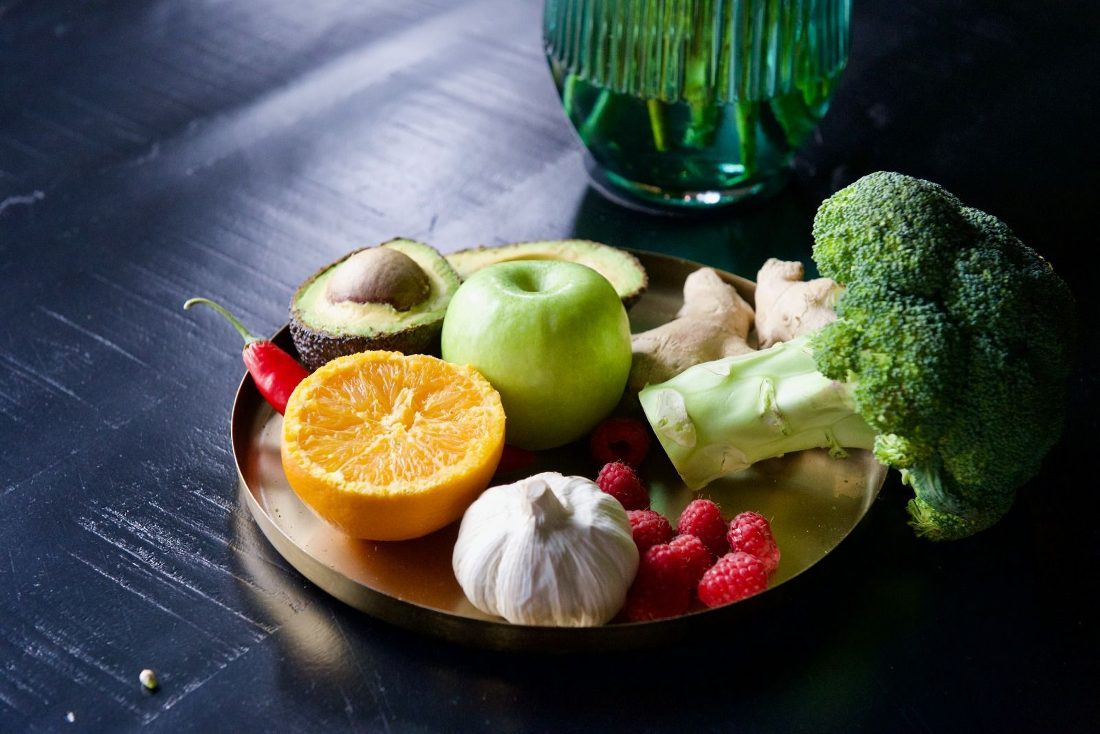 Verse groenten en fruit op een rond bord met een groene fles op een donkere tafel.