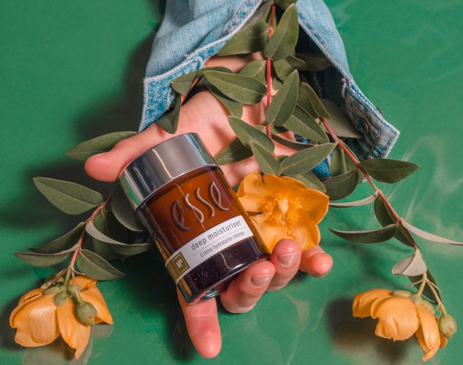 Hand die een klein amberkleurig flesje vasthoudt met oranje bloemen en groene bladeren op een turquoise achtergrond.