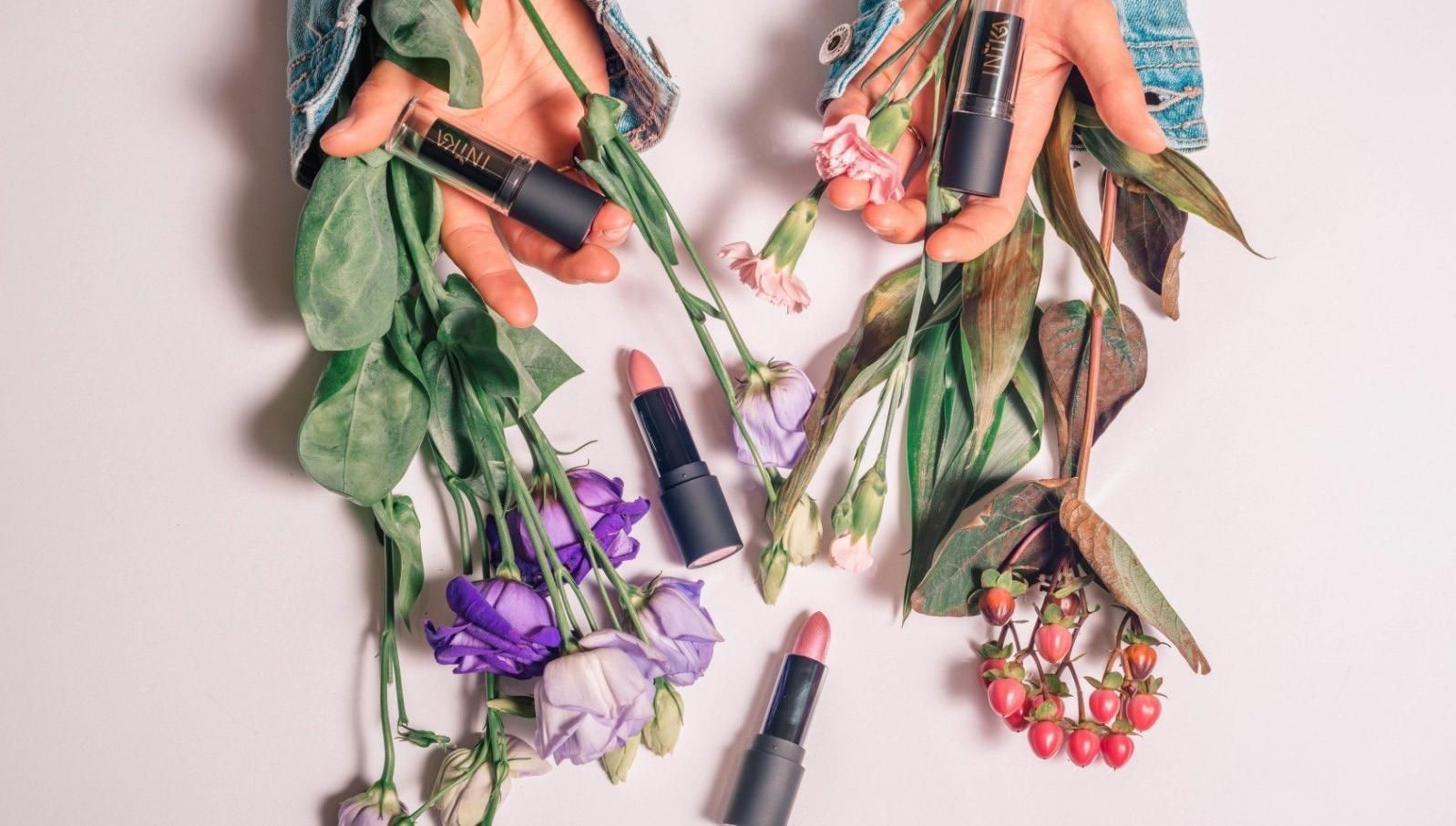 Bloemen en bladeren gerangschikt rondom lippenstiftbuisjes op een lichtroze achtergrond.