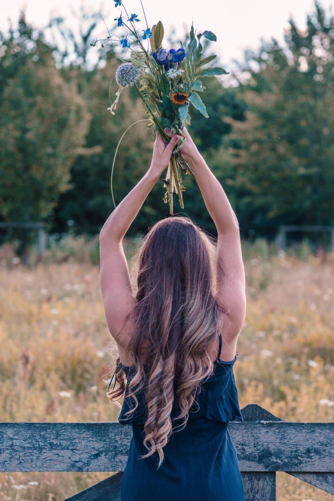 Vrouw in een donkere jurk die een boeket boven haar hoofd houdt in een grasveld, van achteren gezien.