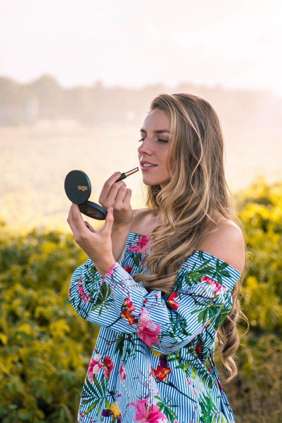 Vrouw brengt buiten make-up aan in een bloemenjurk, met een compact spiegeltje in haar hand, in een zonnig veld.