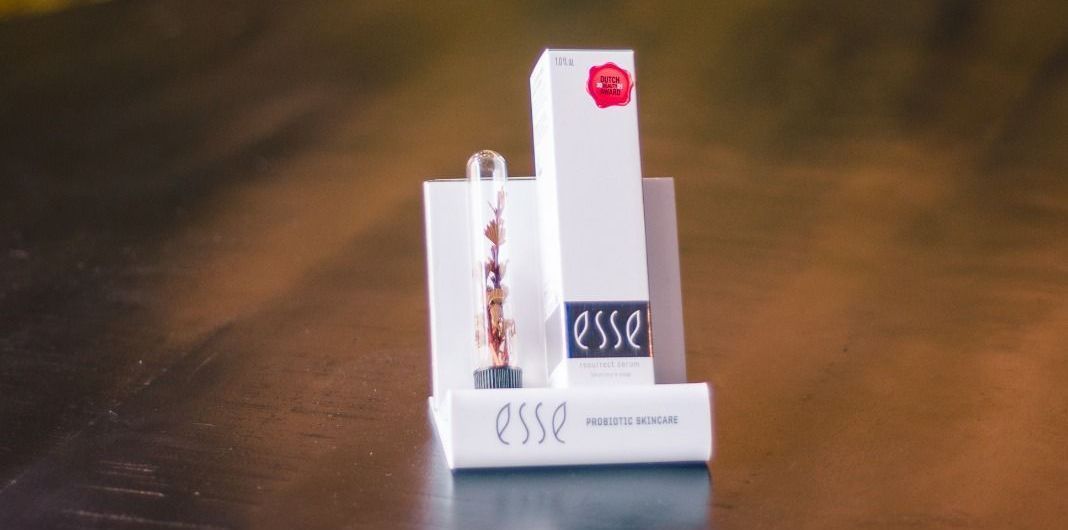 Twee witte cosmeticaverpakkingen met rode en donkere etiketten op een wazige bruine achtergrond.