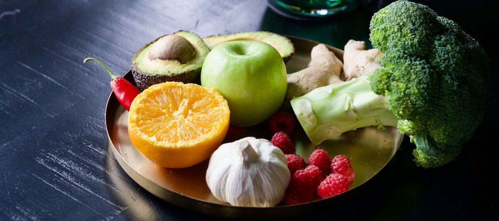 Verse groenten en fruit op een donkere tafel, waaronder broccoli, appel, sinaasappel, knoflook en bessen.