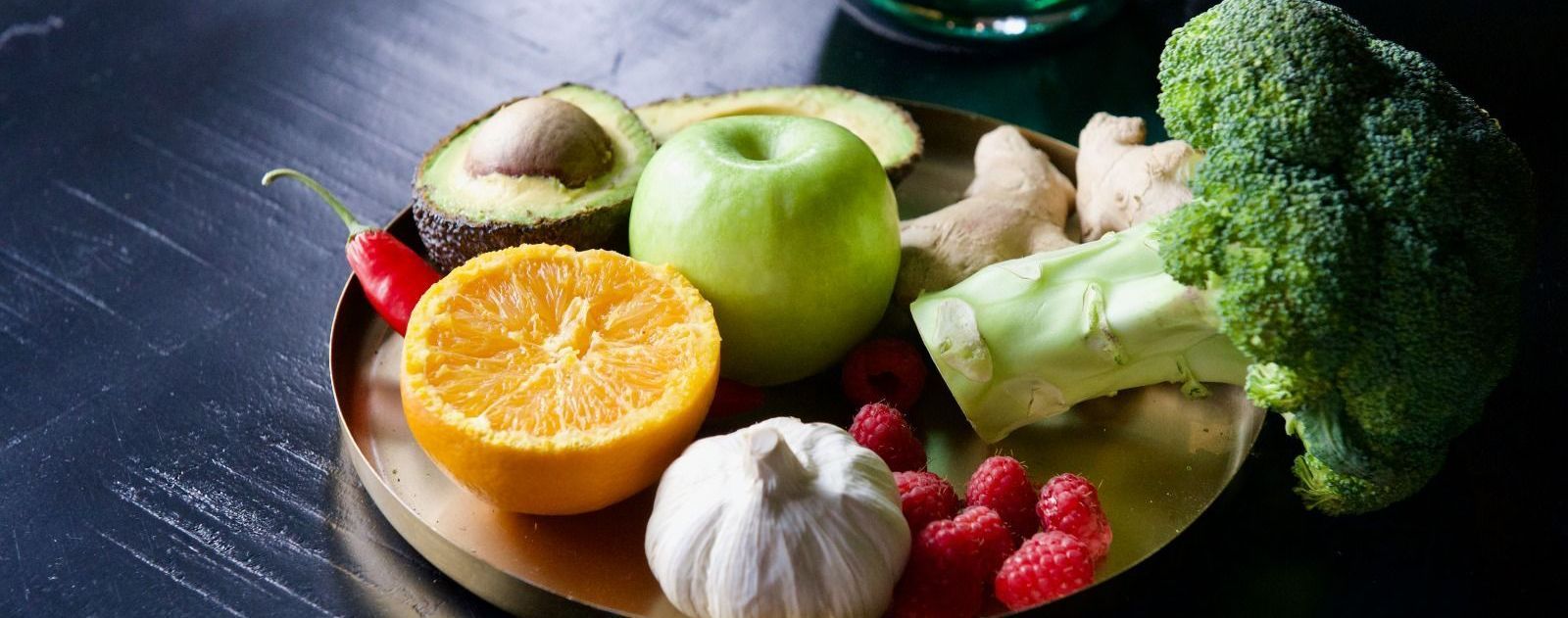 Verse groenten en fruit op een houten plank, waaronder broccoli, knoflook, appel, sinaasappel, avocado, bessen en gember.