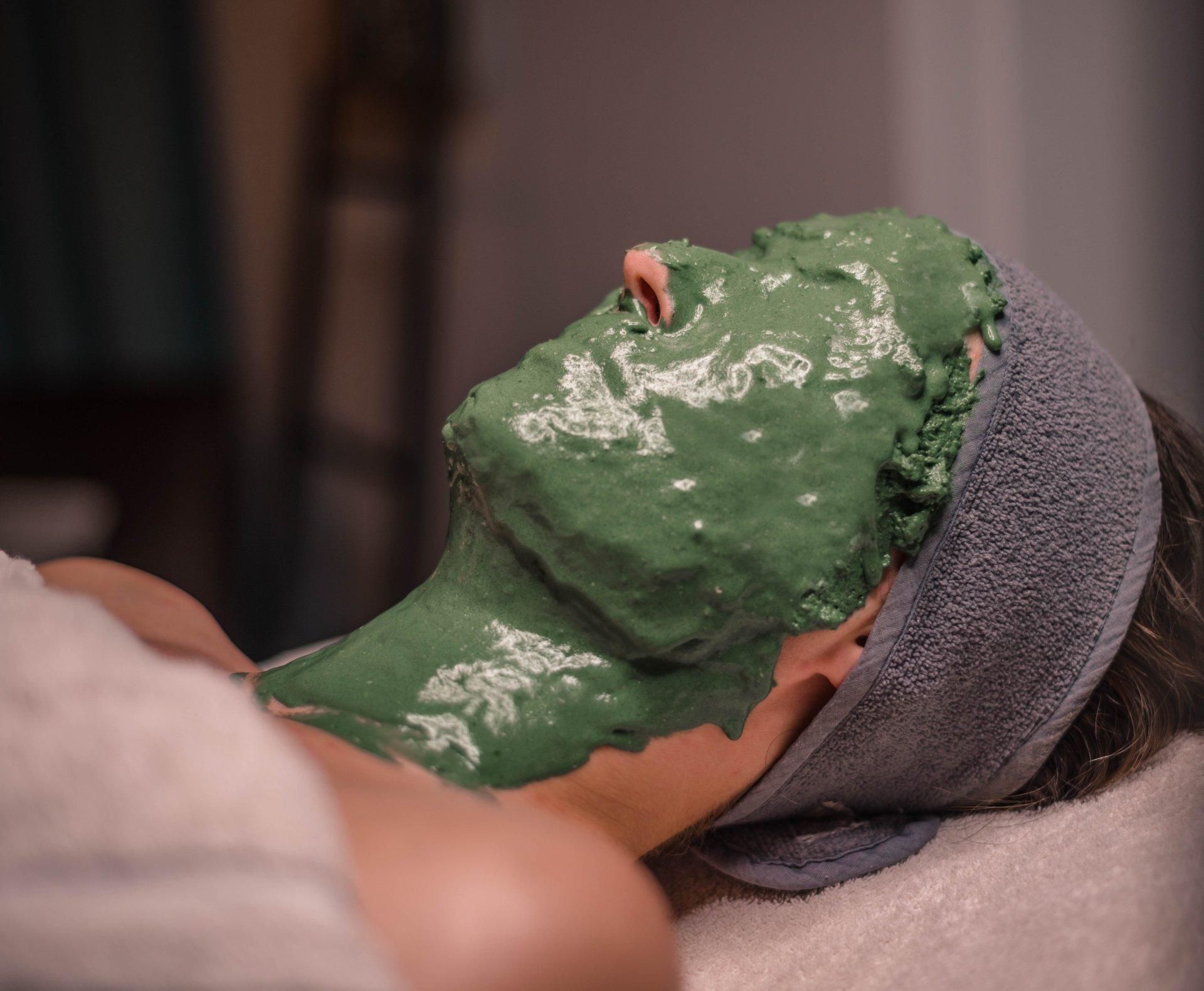 Een persoon ligt op de grond met een groen gezichtsmasker en een handdoek om het hoofd gewikkeld.