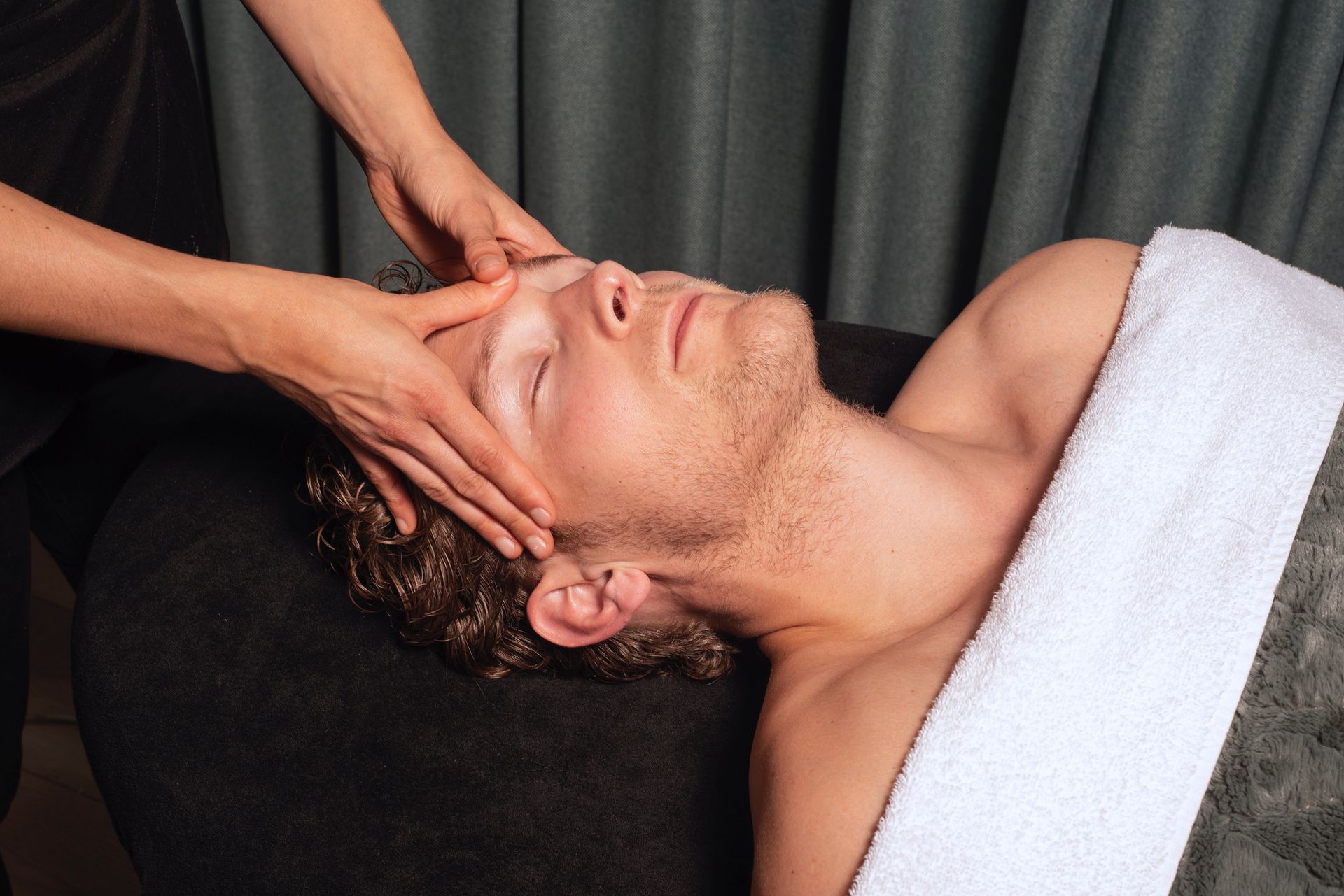 Een massagetherapeut geeft een gezichtsmassage aan een liggende cliënt met een witte handdoek.
