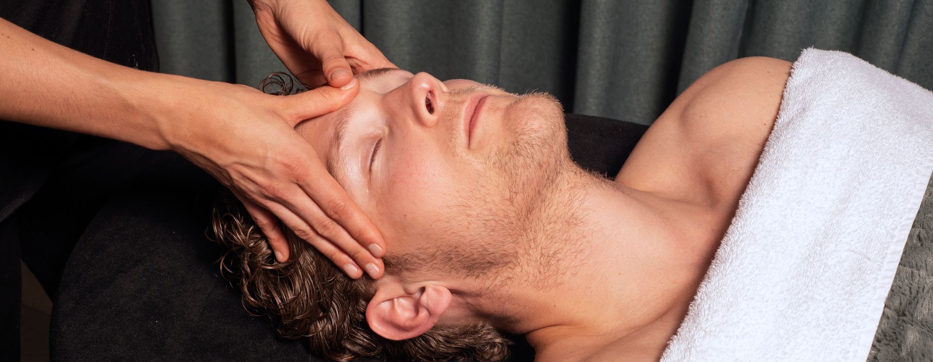 Een massagetherapeut geeft een gezichtsmassage aan een liggende cliënt onder een witte handdoek.
