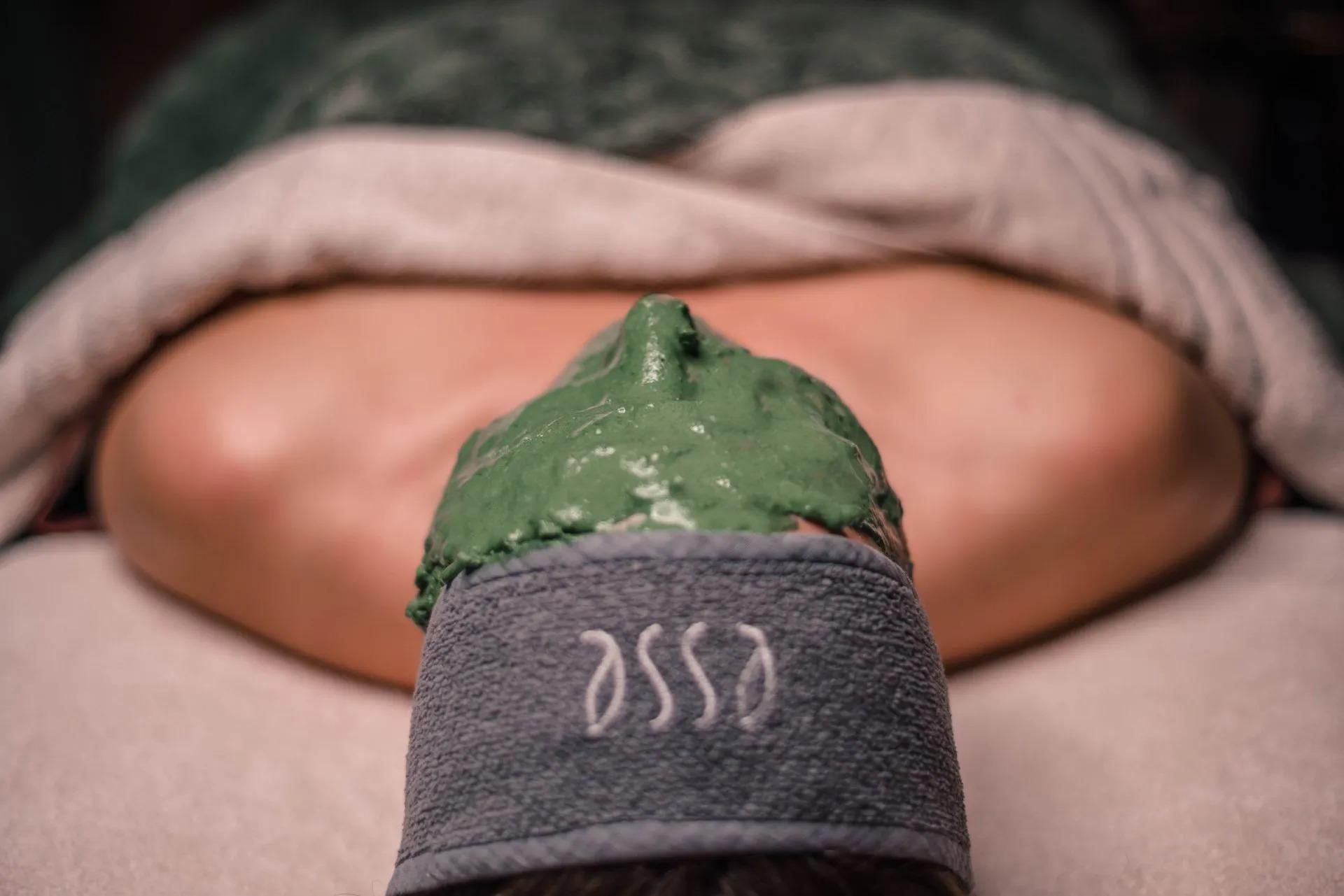 Een persoon ligt op een bed met een groen gezichtsmasker en een handdoek over het haar.