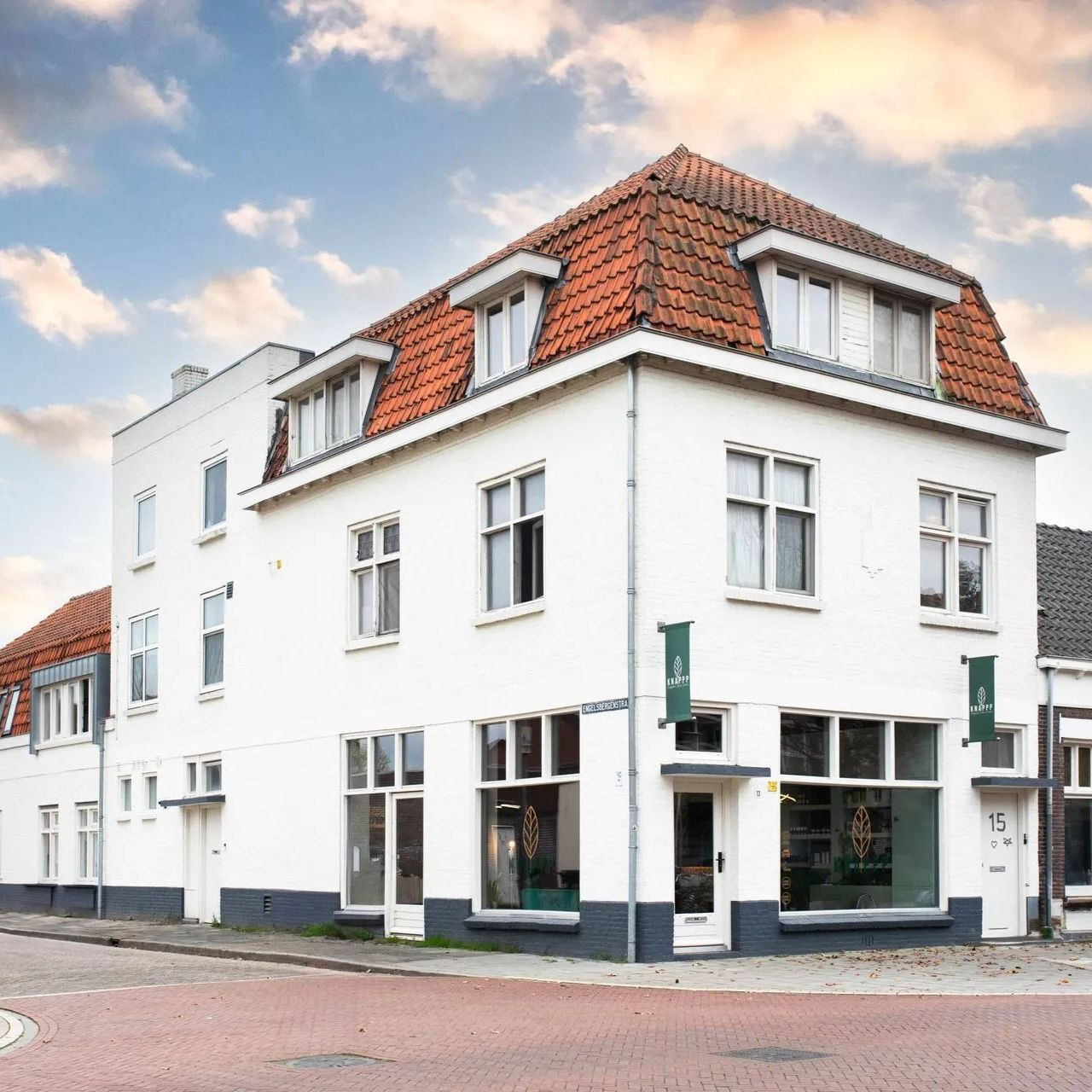 Wit hoekpand met rood pannendak aan een straat, gezien op een heldere dag.
