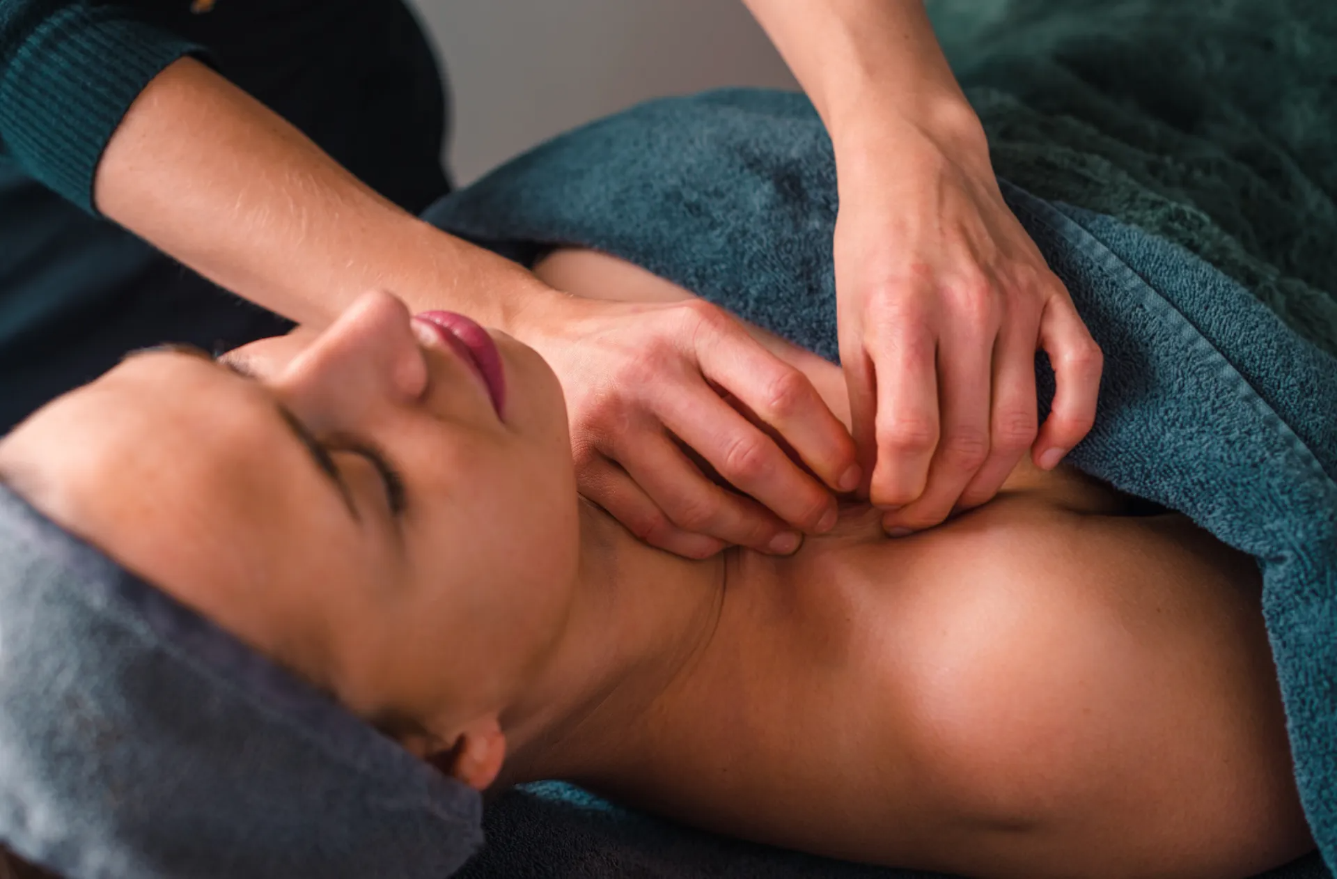 Handen geven een schouder- en nekmassage aan een ontspannen persoon die in een turquoise handdoek is gewikkeld.