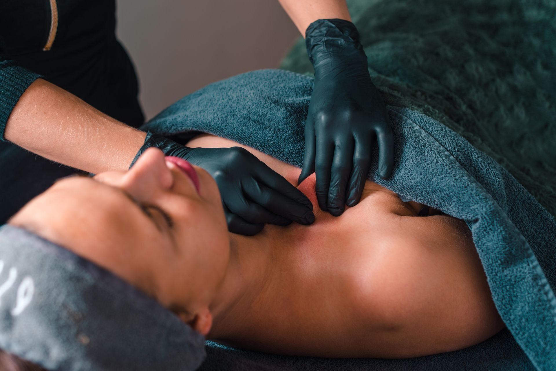 Handen met handschoenen voeren een rugmassage uit op een persoon die op een massagetafel ligt onder een turquoise handdoek.