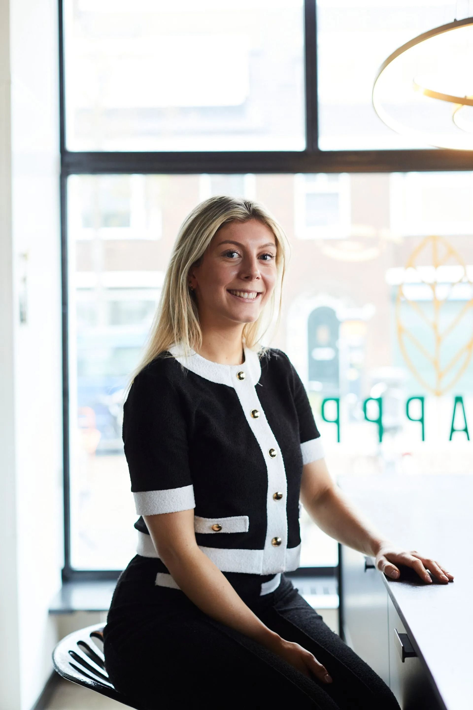 Vrouw in een zwart-witte jurk zit bij een raam en glimlacht achter een bureau in een licht kantoor.