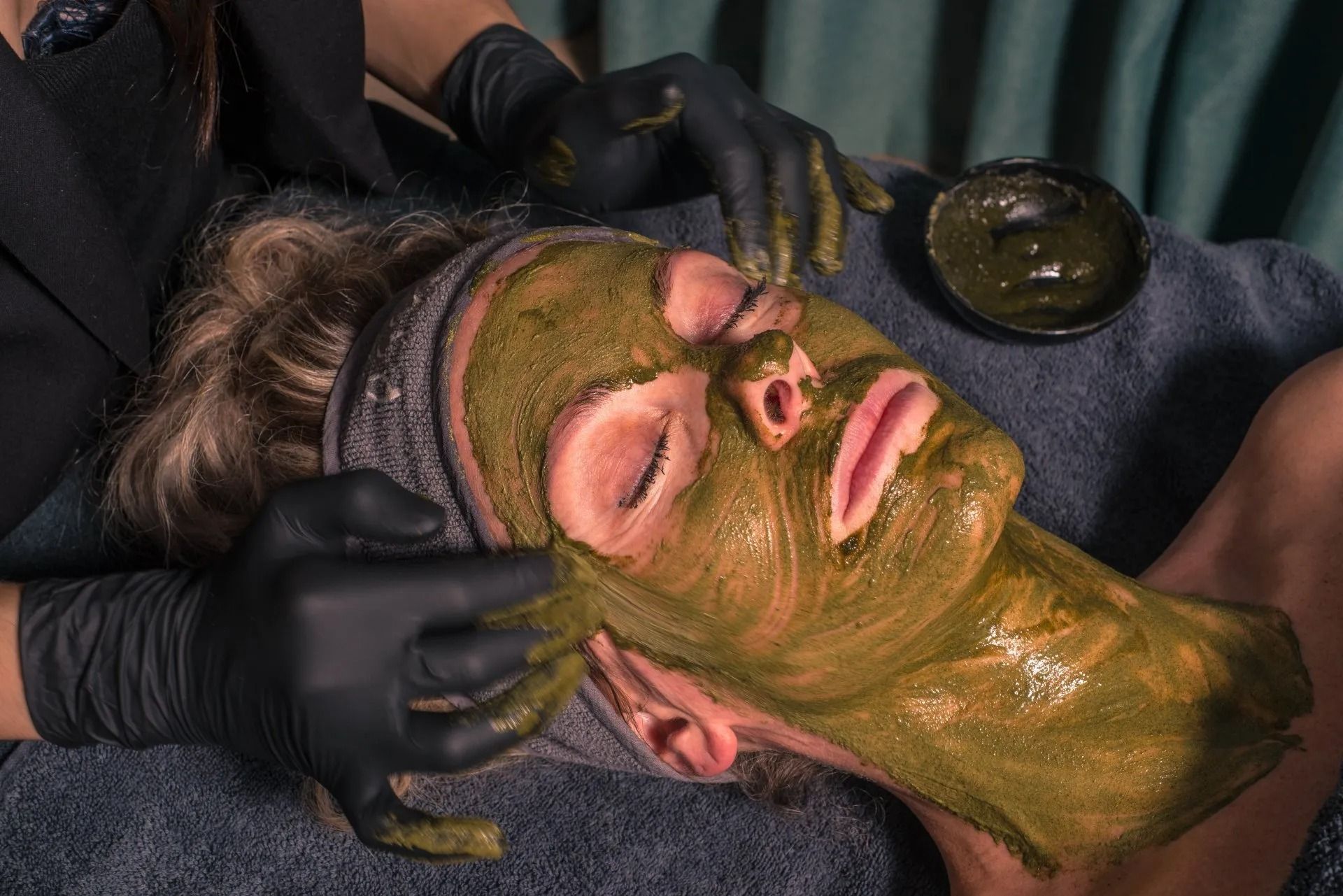Persoon die een groen gezichtsmasker en oogbehandeling krijgt tijdens een gezichtsbehandeling in een spa.