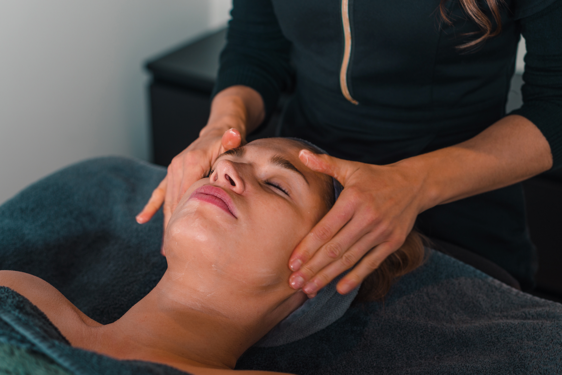 Gezichtsmassage waarbij de handen het gezicht van een vrouw zachtjes vasthouden terwijl ze op een massagetafel ligt.