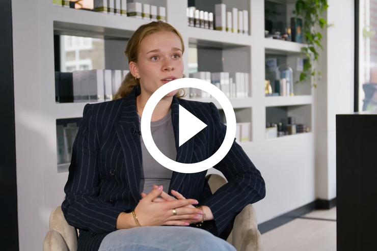 Vrouw zit in een fel verlicht kantoor en spreekt voor de camera, met een afspeelknop op de achtergrond.