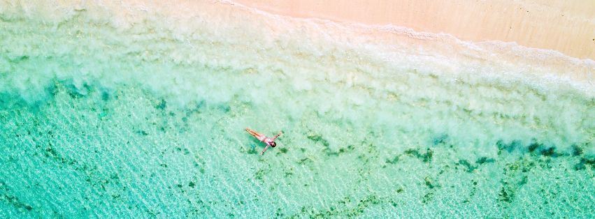 Luchtfoto van een turquoise strand met een eenzame persoon die vlakbij de kust drijft.