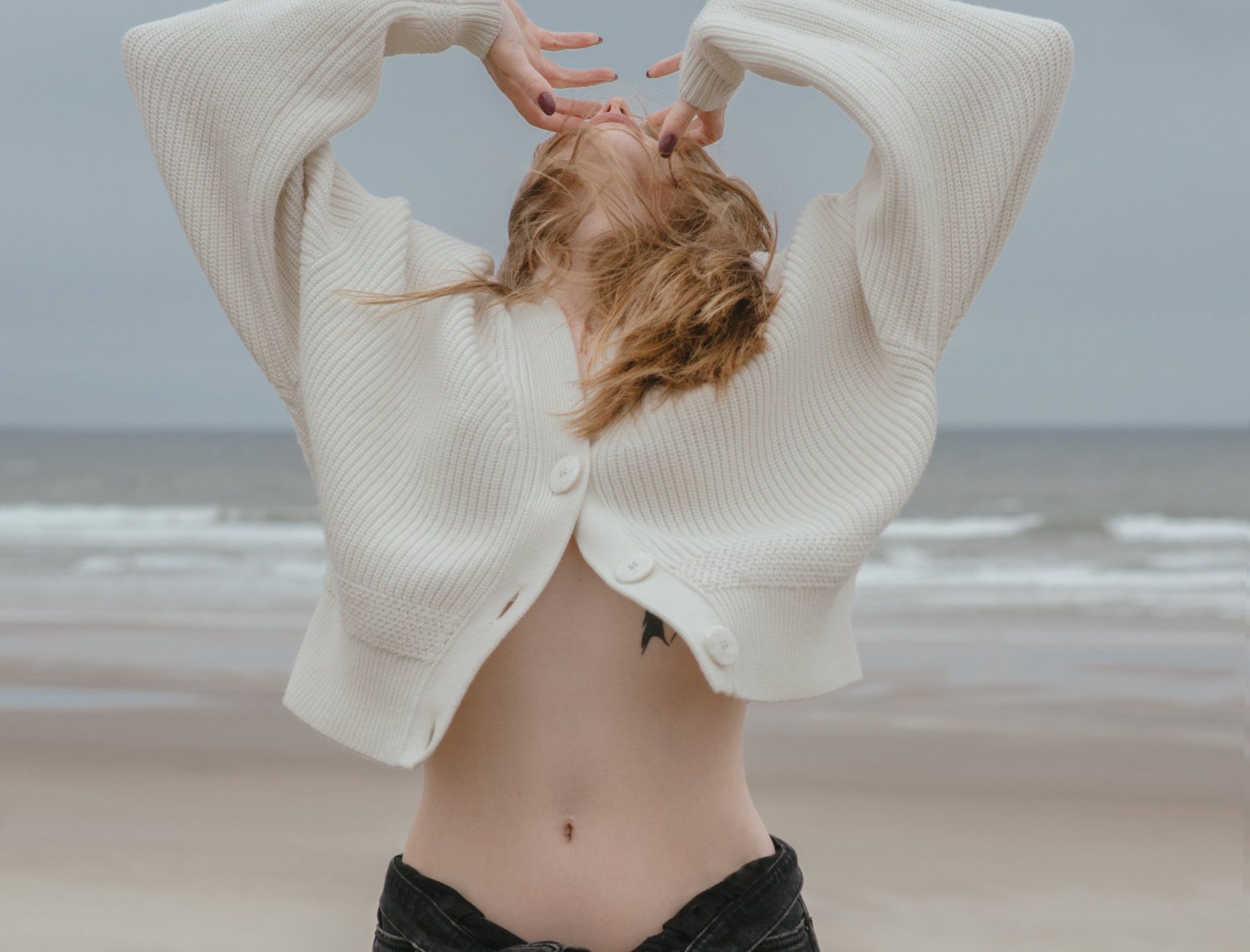 Persoon in een crèmekleurige trui poseert op het strand met de armen omhoog, met oceaangolven op de achtergrond.