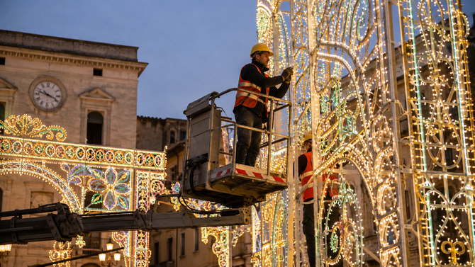 Un operaio su una piattaforma elevatrice installa le luci su una strada addobbata al crepuscolo, con archi illuminati e una torre dell'orologio nelle vicinanze.
