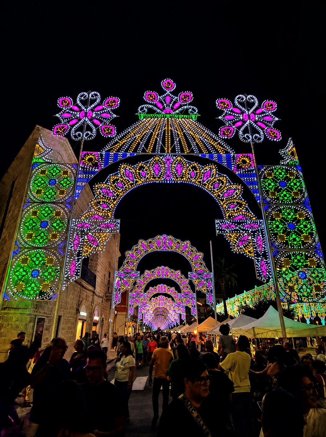 Un arco del festival notturno illuminato da colorati fiori a LED si erge sopra la folla al crepuscolo.