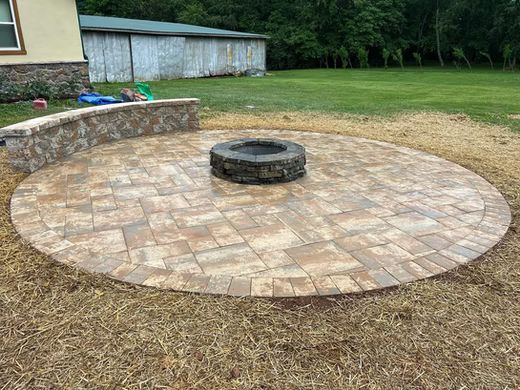 Backyard patio with circular stone paver area and central fire pit, bordered by dry grass and a low retaining wall