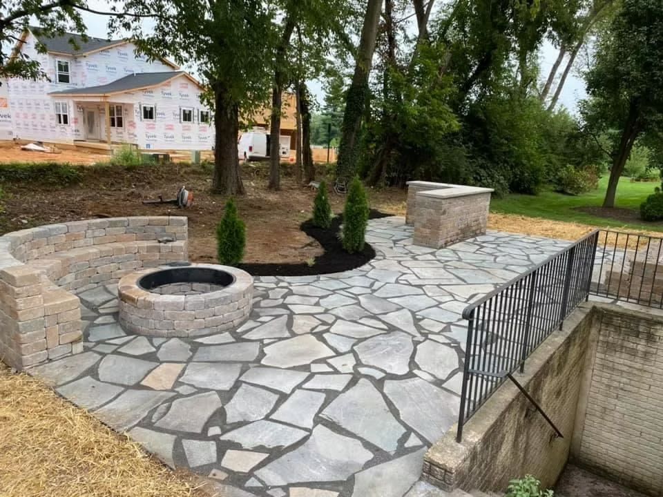 Stone patio with circular fire pit, built-in bench, and black railing in a backyard surrounded by trees